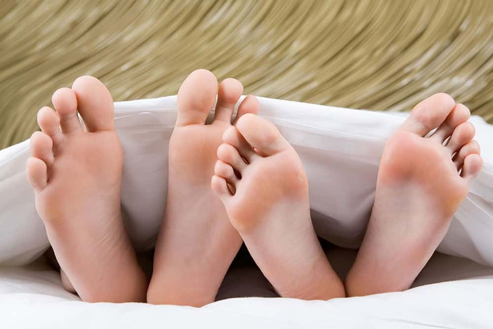 Couple's feet peeking out from under a blanket, illustrating shared living and potential foot bacteria transfer, relevant to toenail health and hygiene.
