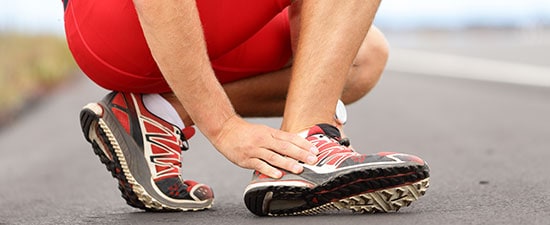 Runner experiencing foot pain while holding ankle, wearing red athletic shorts and running shoes on a track surface, relevant to conditions causing pain on the outside of the foot.