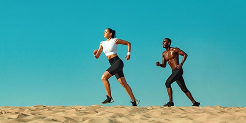 Two runners on a sandy beach, one female in a white crop top and black shorts, and one male shirtless in black shorts, illustrating the importance of foot health and common running injuries.
