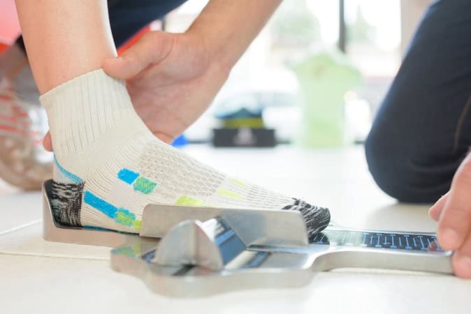 Person measuring foot size using a Brannock device while wearing a sock, emphasizing proper shoe fitting for foot health.