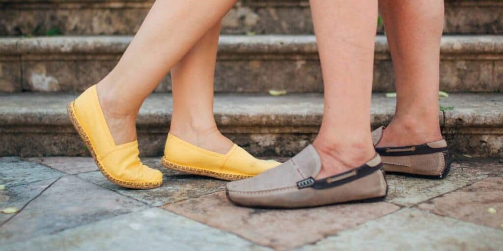 Couple wearing casual summer shoes, one in yellow espadrilles and the other in brown slip-ons, on outdoor stone steps, highlighting comfortable footwear choices for foot health.