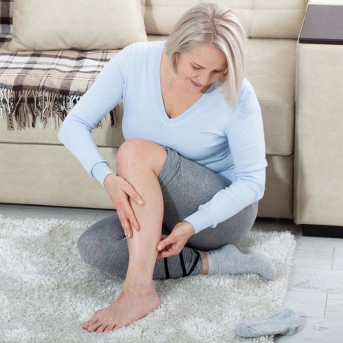 Older woman experiencing leg pain, sitting on the floor and holding her calf, indicating discomfort or injury.
