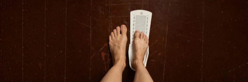 Person standing on a white bathroom scale with bare feet, measuring weight on a dark wooden floor.